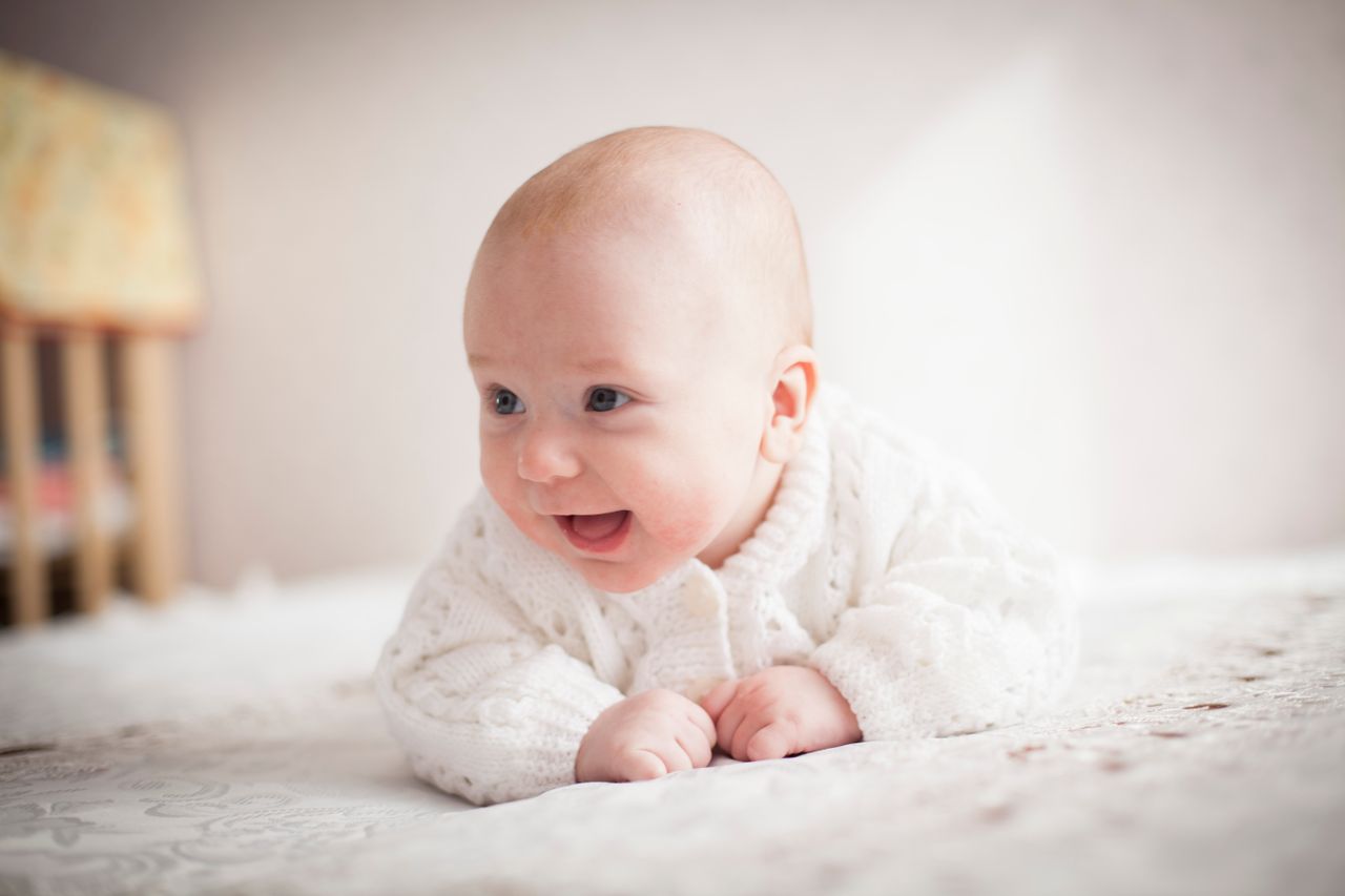 about-us-02 A cute baby smiling on a soft, patterned blanket indoors. Bright and heartwarming moment.