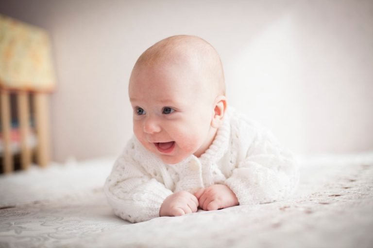 A cute baby smiling on a soft, patterned blanket indoors. Bright and heartwarming moment.